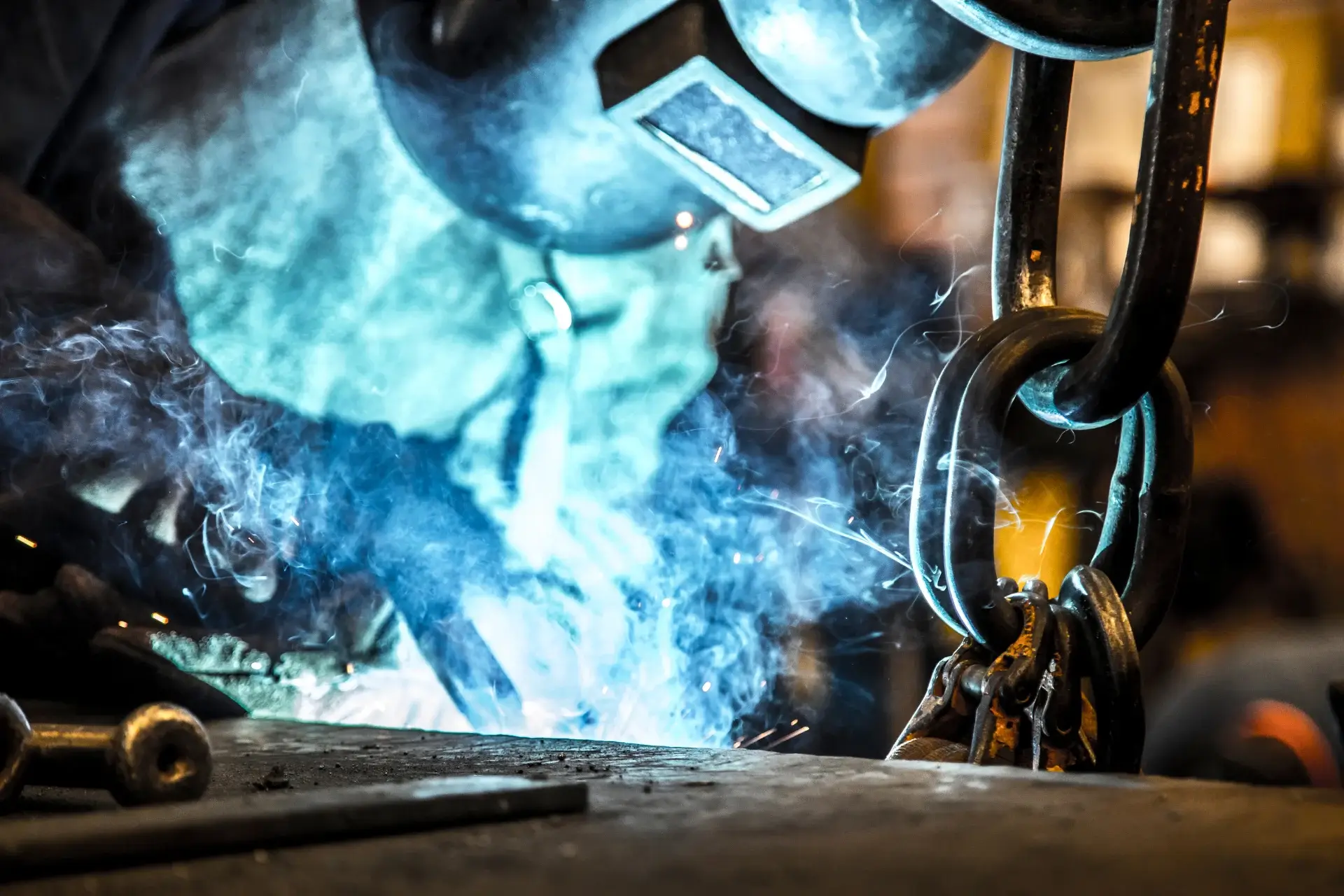 Welder working on a steel component, with sparks and smoke beside heavy lifting chains.